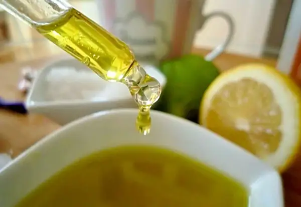 Tropfen eines goldenen Öls fallen aus einer Pipette in eine Schale. Zitronen und Limetten im Hintergrund deuten auf natürliche Inhaltsstoffe für Kosmetik hin, wie z.B. Zitronensäure.