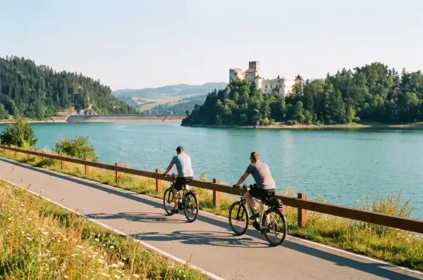 Dwóch rowerzystów na ścieżce Velo Dunajec Czorsztyn, zamek w tle i turkusowa tafla jeziora.