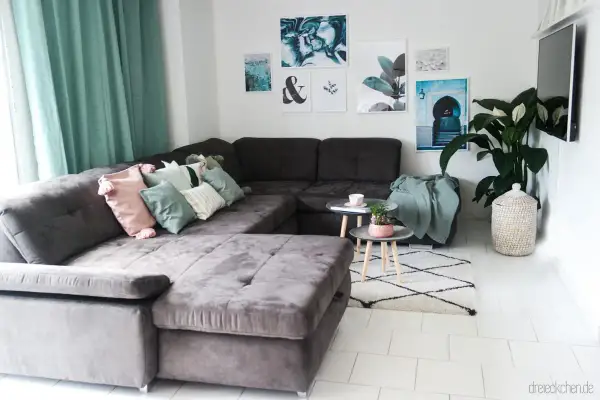 Gemütliches wohnzimmer einrichten mit einer großen, dunklen Couch, vielen Kissen in Pastelltönen und mintgrünen Akzenten. Bilder und eine Pflanze schmücken die Wand.