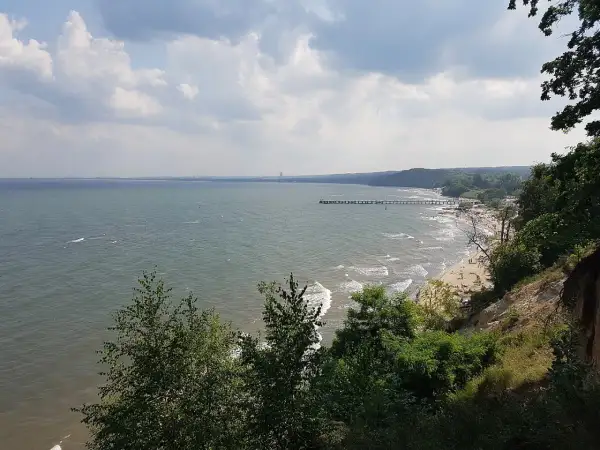 Widok na morze i plażę z klifu orłowskiego. W oddali widać molo i ludzi odpoczywających na piasku.