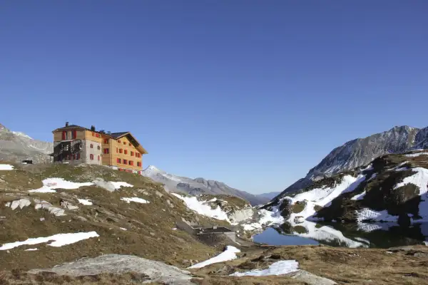 Das Pfitscher Joch Haus thront auf einem Hügel neben einem Bergsee. Die Alpenlandschaft ist von Schnee und braunem Gras bedeckt.