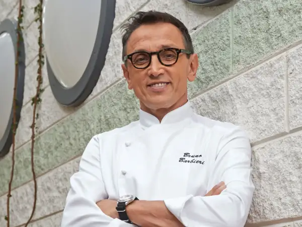 Bruno Barbieri, chef stellato Michelin, sorride con le braccia conserte davanti a un muro di pietra.