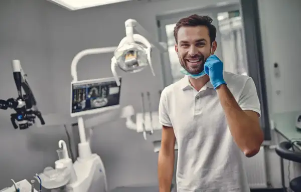 Uśmiechnięty dentysta w gabinecie, gotowy do pracy. Zastanawiasz się, ile trwa plombowanie zęba?