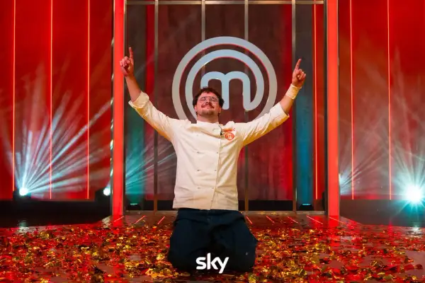 Vincitore del finale MasterChef 2026 in ginocchio tra coriandoli dorati, con le braccia alzate e il logo di MasterChef sullo sfondo.