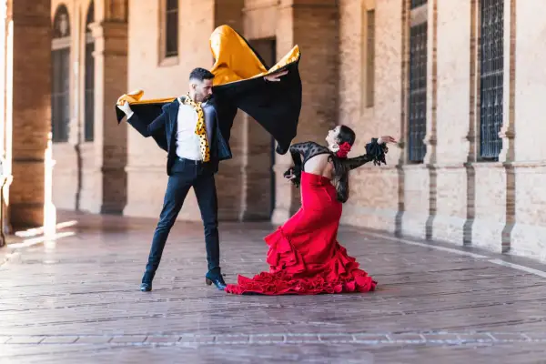 Pareja bailando flamenco, él con capa negra y amarilla, ella con vestido rojo voluminoso. La imagen evoca la diferencia entre flamenco y sevillanas.