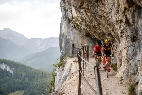 Dwóch rowerzystów pokonuje malownicze trasy rowerowe w Austrii, jadąc wąską ścieżką przy skalnej ścianie z widokiem na góry.