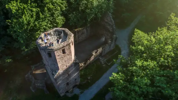 Blick von oben auf die Ruine der Barbara Kapelle, umgeben von grünen Bäumen. Eine Gruppe von Menschen genießt die Aussicht vom Turm.