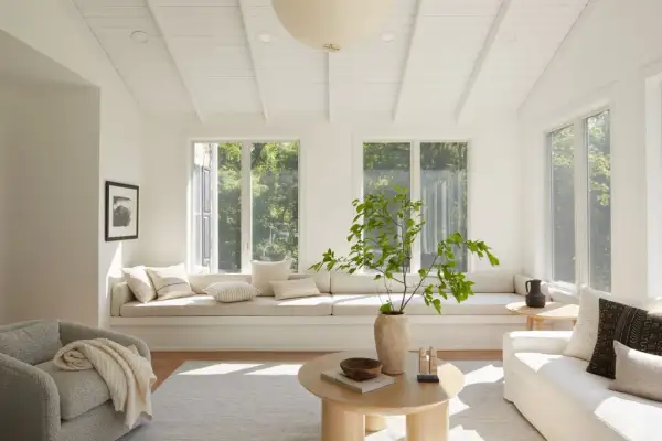 A bright living room with neutral interior design, featuring a large window seat, a textured armchair, and a light wood coffee table.
