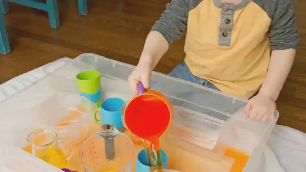 A child enjoys Montessori water play, pouring orange liquid from a colorful pitcher into a blue cup.