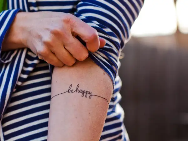 Un brazo con una camiseta a rayas azules y blancas muestra un delicado tatuaje de caligrafía que dice "be happy", una forma de arte para transformar cicatrices.