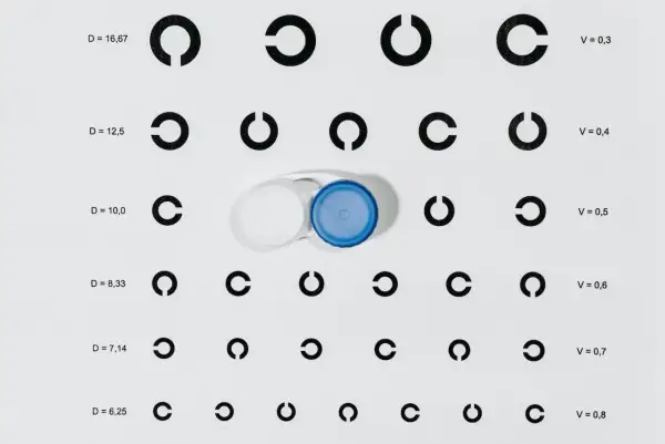 Tabela optometryczna z cyframi i otwartymi okręgami, obok pojemnik na soczewki kontaktowe.