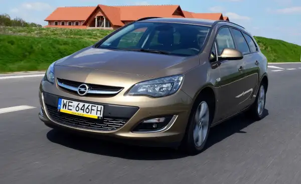 Brązowy Opel Astra J automat w wersji kombi jedzie po drodze. W tle widać domy z czerwonymi dachami.
