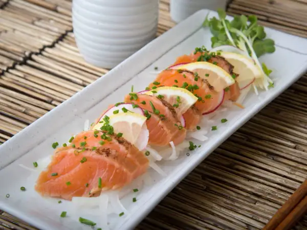Tataki de salmón fresco, finamente cortado y adornado con rodajas de limón, rábano y cebollino, servido sobre una cama de finas hebras de cebolla.