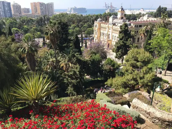 Vista panorámica de un jardín exuberante con flores rojas y palmeras, un edificio histórico y el mar al fondo en Málaga.
