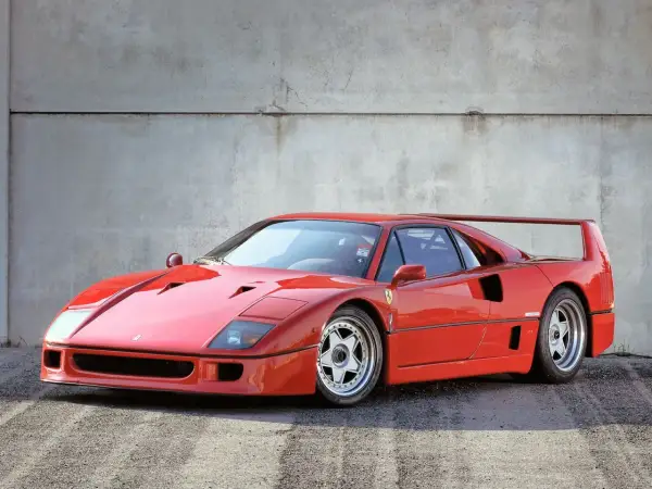 Ikoniczny czerwony Ferrari F40 z charakterystycznym skrzydłem, zaparkowany na tle betonowej ściany.