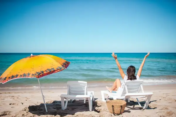 Kobieta cieszy się słońcem na plaży, zastanawiając się, ile dni urlopu jeszcze jej zostało. Obok parasol i leżaki.