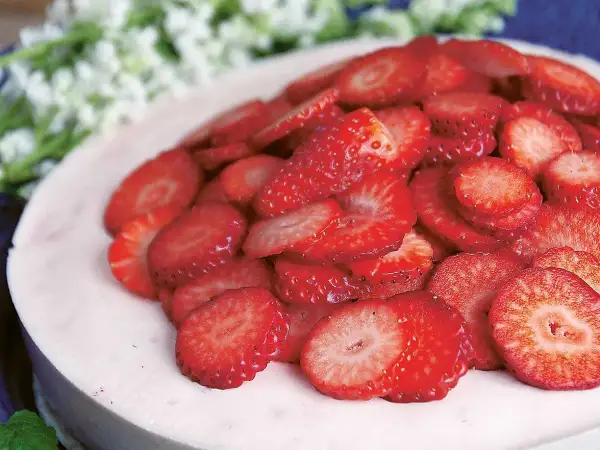En läcker jordgubbsmousse tårta, toppad med färska jordgubbsskivor och vita blommor i bakgrunden.