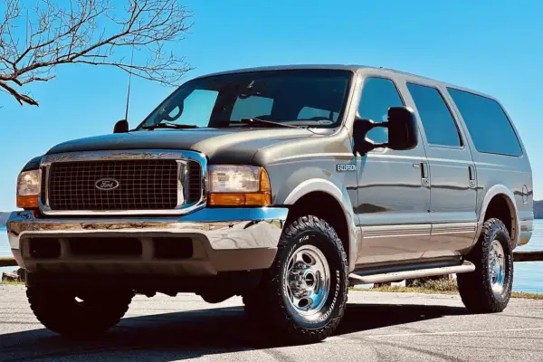 Brązowy Ford Excursion z chromowanymi dodatkami i terenowymi oponami stoi na tle błękitnego nieba i nagich drzew.