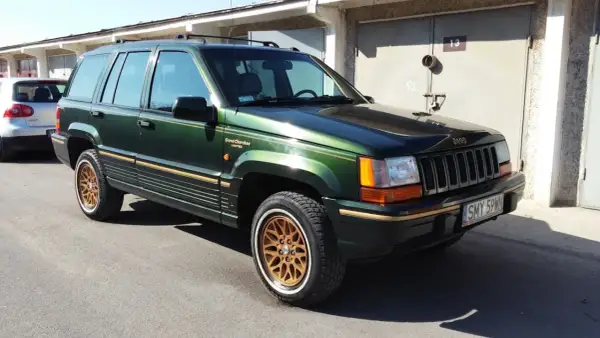 Zielony Jeep Grand Cherokee Limited z lat 90. na złotych felgach, zaparkowany przed garażami.