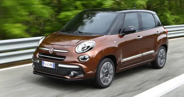 Brązowy Fiat 500L z czarnym dachem pędzi po drodze, otoczony zielenią.