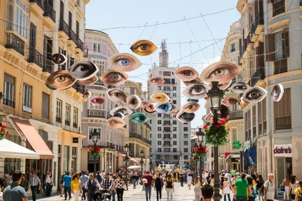 Ojos flotantes decoran una calle famosa de Málaga, observando a la multitud que pasea entre edificios históricos.