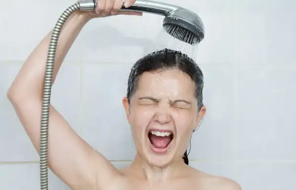 Une femme crie sous une douche froide, visiblement contrariée par un problème d'eau chaude.