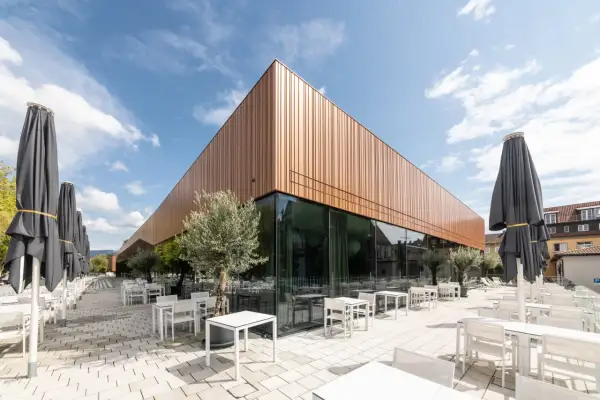 Moderne Architektur der Inselhalle Lindau mit kupferfarbener Fassade, Glasfront und vielen leeren Tischen und Stühlen auf einer gepflasterten Terrasse.