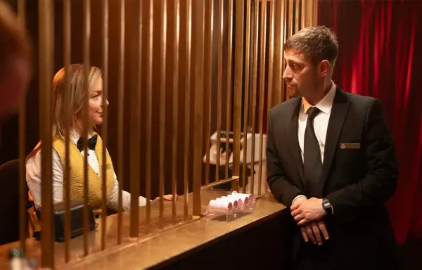 En kvinna i gul väst och fluga samt en man i kostym möts bakom en disk. En scen från en ny brittisk serie på SVT.