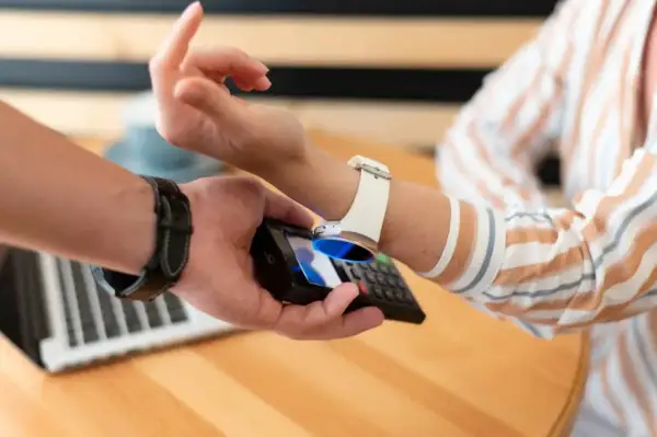 Zbliżenie na dłoń trzymającą telefon, który zbliża się do białego zegarka na ręce. Pokazuje, jak naładować zegarek telefonem.