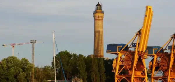 Wysoka, żółta latarnia morska w Gąskach góruje nad portem, obok potężnych, żółtych dźwigów portowych i zielonych drzew.