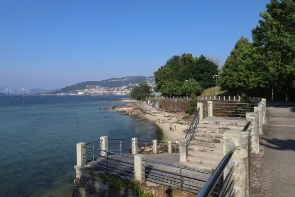 Vista de la playa de las barcas con gente disfrutando del sol y el mar. Al fondo, un puente y una ciudad en la ladera de una montaña.