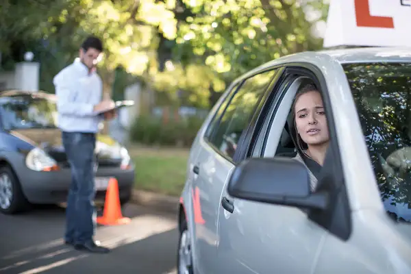 Joven nerviosa al volante, con un instructor anotando. ¿Se puede sacar el carnet de conducir sin autoescuela?