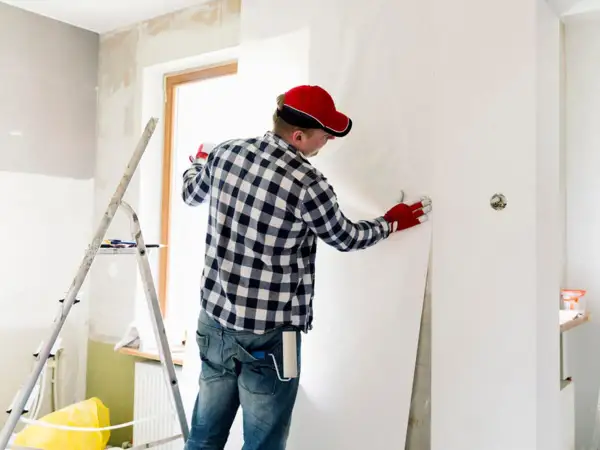 Mann mit roter Kappe und Handschuhen kleistert Vliestapete ein, um eine Wand zu tapezieren.