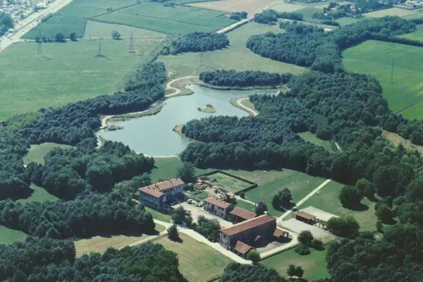 Un'oasi di pace: un lago serpeggia tra fitti alberi, creando un bosco in città. Edifici rurali si affacciano su prati verdi.