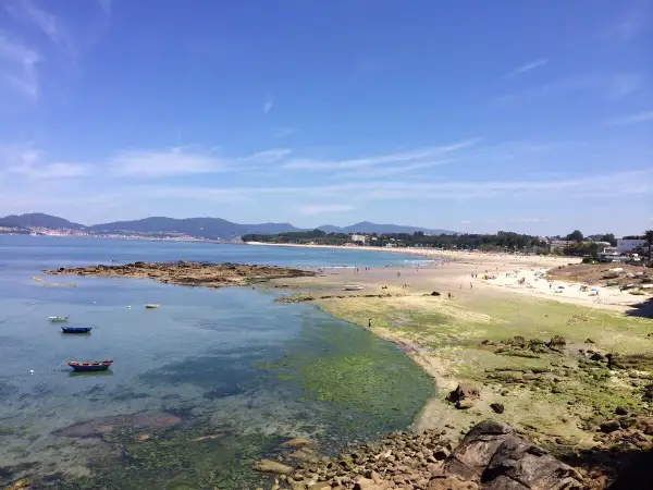 Vista de la playa de A Calzoa en Vigo, con gente disfrutando del sol y el mar. Barcas pequeñas flotan en aguas cristalinas.