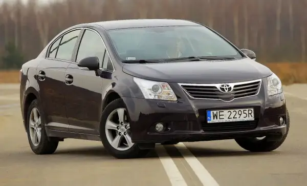 Ciemny sedan Toyota Avensis na drodze. Kierowca za kierownicą. Szczegółowe dane techniczne tego modelu są dostępne online.