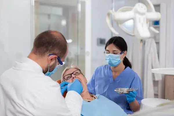 Dentysta zakłada białe plomby na NFZ starszej pacjentce. Asystentka stomatologiczna pomaga.