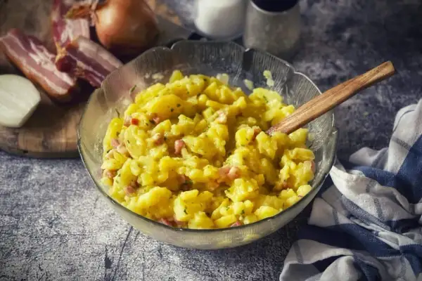 Ein dampfender, warmer Kartoffelsalat mit Speck und Zwiebeln, serviert in einer Glasschüssel mit Holzlöffel.