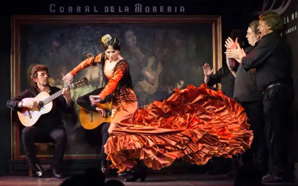Una bailaora de flamenco, con un vestido naranja voluminoso, baila al son de guitarras. Es un espectáculo de flamenco vibrante.