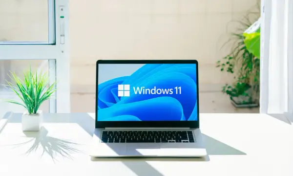 Laptop z logo Windows 11, gotowy na aktualizację 25H2. Na biurku obok rośliny, w jasnym pomieszczeniu.