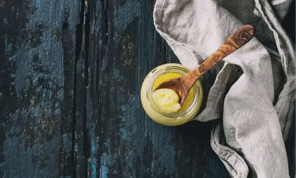 Ein Glas mit goldgelbem Ghee, daneben ein Holzlöffel mit einer Portion. Ghee ist geklärte Butter, ein vielseitiges Kochfett.