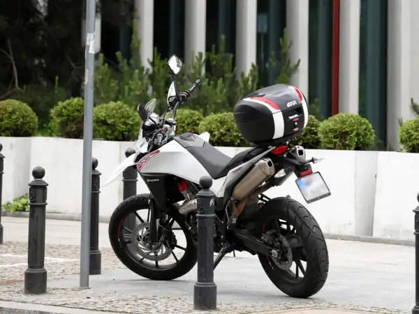 Biały motocykl z czarnym kufrem Givi zaparkowany na chodniku, obok słupków.
