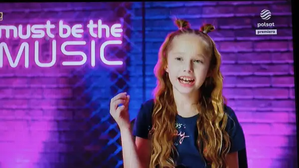 Lili chce śpiewać autor. Dziewczynka z burzą rudych loków i dwoma kucykami na głowie, na tle neonowego napisu "must be the MUSIC".