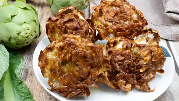 Piatto di carciofi alla giudia dorati e croccanti, con carciofi freschi sullo sfondo.