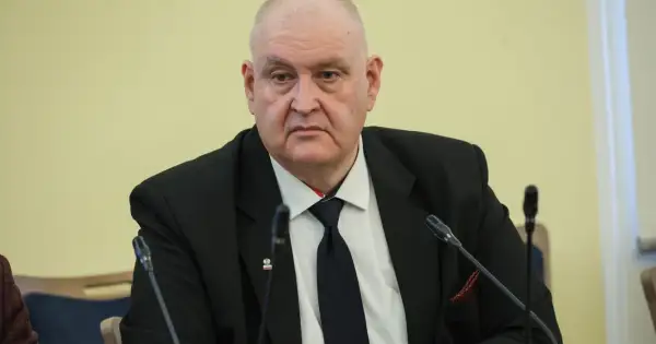 Prezes Trybunału Konstytucyjnego w garniturze, z przypinką z godłem Polski, siedzi przy stole z mikrofonami.