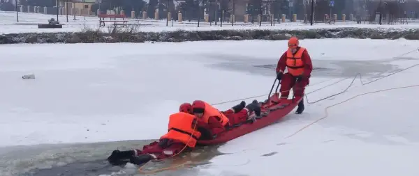Ratownicy w akcji, wyciągają osobę z lodowatej wody za pomocą **sanki ratownicze**. Zimowa sceneria i profesjonalny sprzęt.