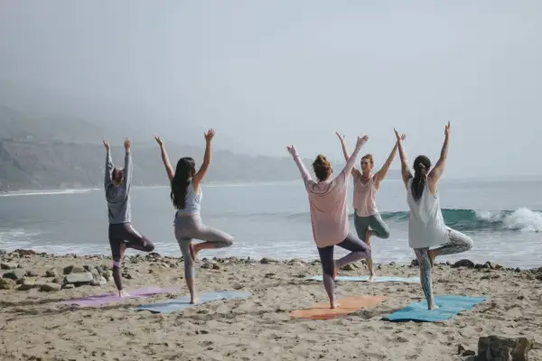 Cinco mujeres practican poses de yoga para 4 en la playa, con los brazos levantados hacia el cielo.