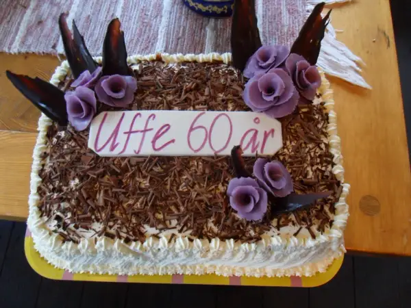 En tårtbotten i långpanna dekorerad med chokladspån, lila rosor och texten "Uffe 60 år".