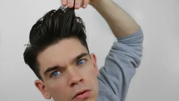 Mann mit einem messy quiff, der sich die Haare hochzieht. Blaue Augen, graues Sweatshirt.