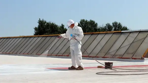 Pracownik w białym kombinezonie natryskuje nowoczesne materiały na dach płaski EPDM, porównując je z papą termozgrzewalną.